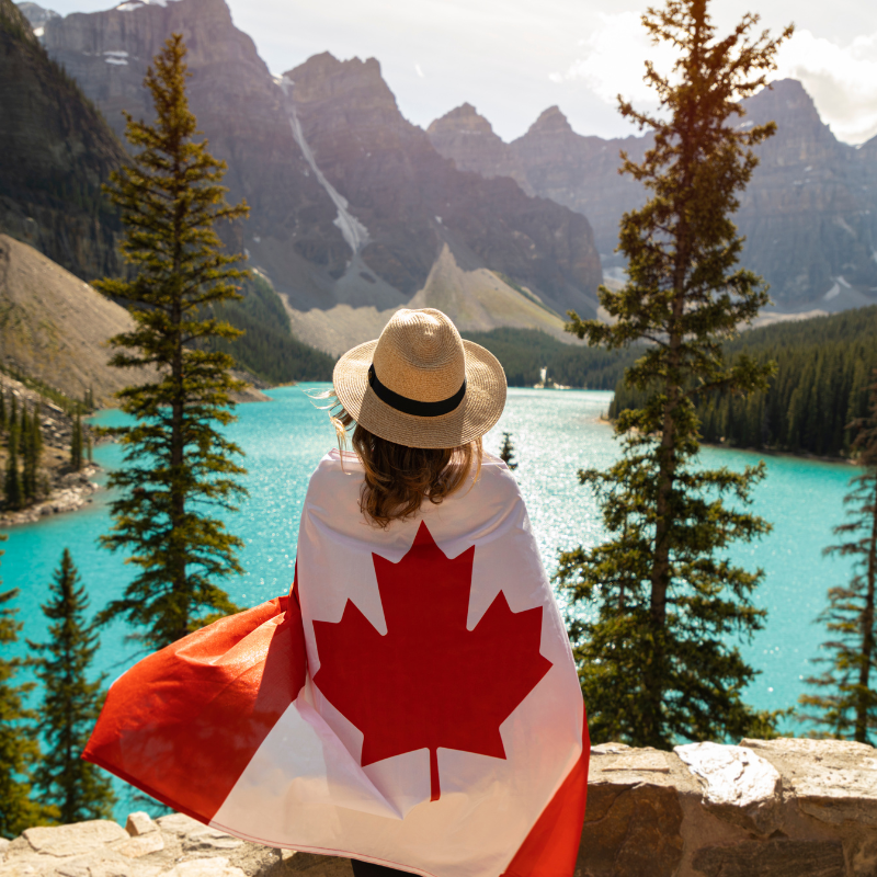 Stock image of a women standing near a Canadian Lake, Looking at the mountains with a Canadian Flag wrapped around her back.