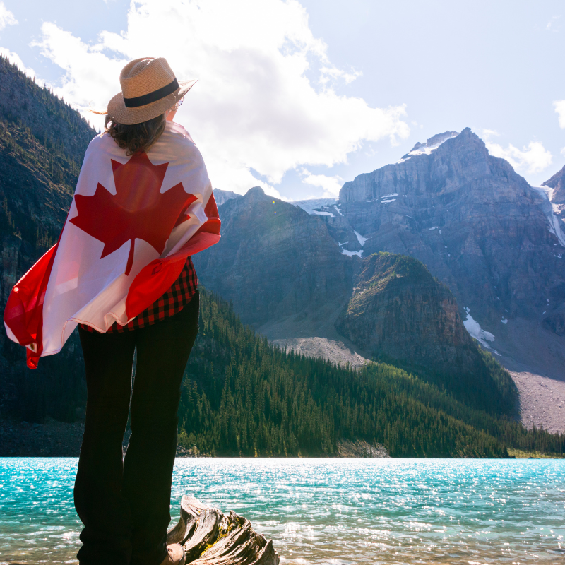 Stock image of a women standing near a Canadian Lake, Looking at the mountains with a Canadian Flag wrapped around her back.