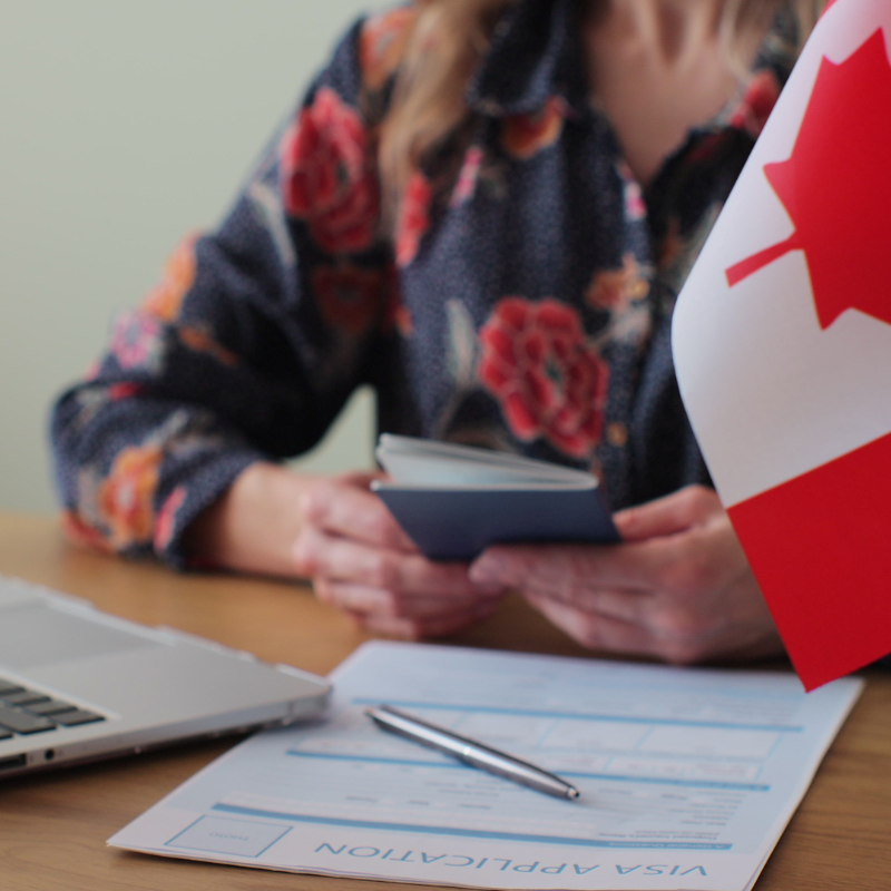 Stock image with a document with pen on it, partial laptop and partial Canadian Flag is visible, women in background (face not visible) holding a passport. Image chosen to demonstrate Canadian work permit specialist IRCC office and client.