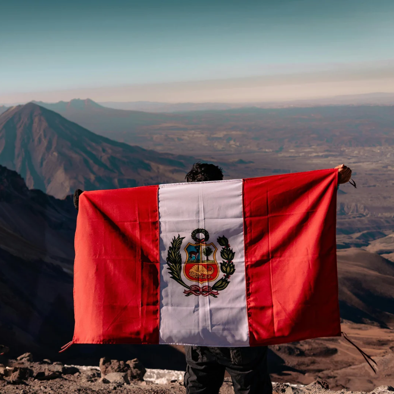 A man standing on top of a mountain holding Peru flag around his shoulder, used to build relevance to the service page topic of Peru Free Trade Agreement Work Permit.
